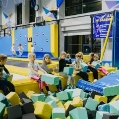 Dzieci siedzą na żółtej ściance w basenie z gąbkami, trzymając kostki. Jedna dziewczyna w niebieskiej bluzie patrzy na nie, jakby to była jakaś baba jaga patrzy zabawa.