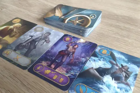 Na stole leżą karty z gry ryzykanci. Widoczny jest stos kart z kompasem, a obok cztery karty przedstawiające postacie: starszego pana z fajką, mężczyznę z aparatem, kobietę z pochodnią i mężczyznę w łodzi na rwącej rzece.
