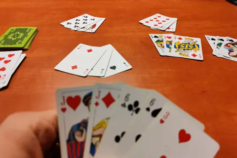 Gracz trzyma karty do gry w remika: walet serce, walet karo, szóstka trefl, szóstka pik, trójka serce. Na stole leżą inne karty.