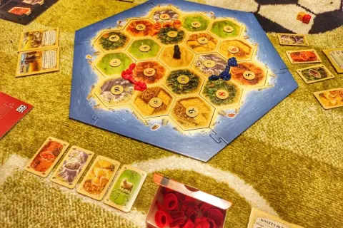 Plansza Catan: Świt Ludzkości, karty surowców i pionki. Rozgrywka w toku, gracze budują osady i drogi.