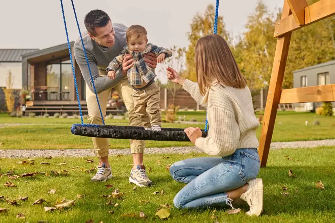 Rodzina cieszy się wspólnymi zabawami na podwórku. Tata trzyma dziecko na huśtawce, a mama zachęca do zabawy.